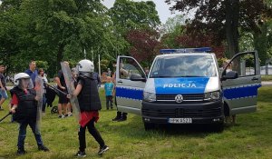 Na zdjęciu widoczny jest w tle radiowóz oznakowany VW T6. Na pierwszym planie widać dzieci przebrane za policjantów OPP, które udają, że walczą ze sobą